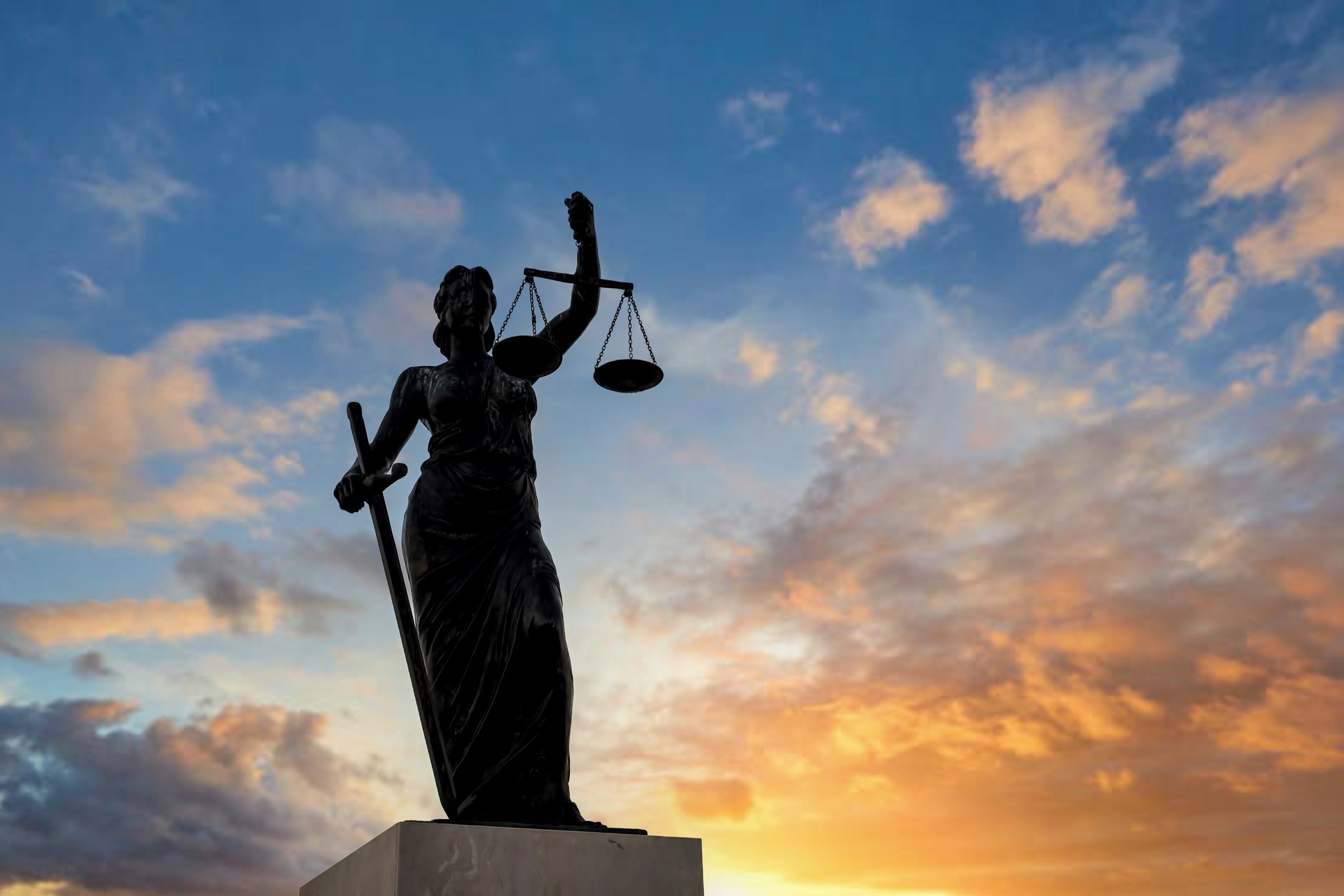 Imagem da estátua da justiça, com um céu azul e alaranjado ao fundo e o sol se pondo.
