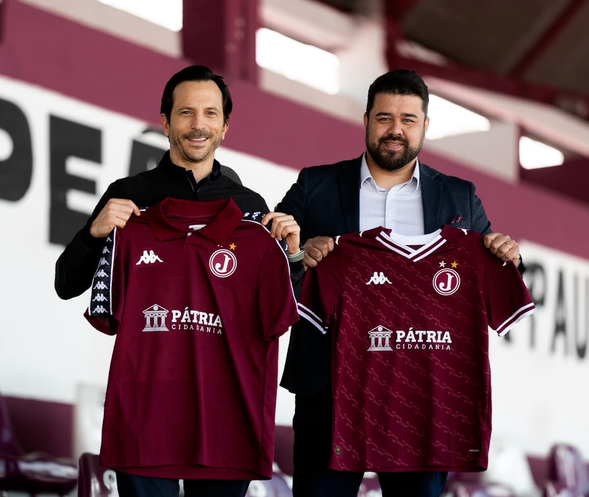 Foto de Vinícius Gama, sócio-fundador da Pátria Cidadania, com Fábio Wolff, sócio-diretor da Wolff Sports. Ambos seguram a icônica camisa grená do Juventus da Mooca.