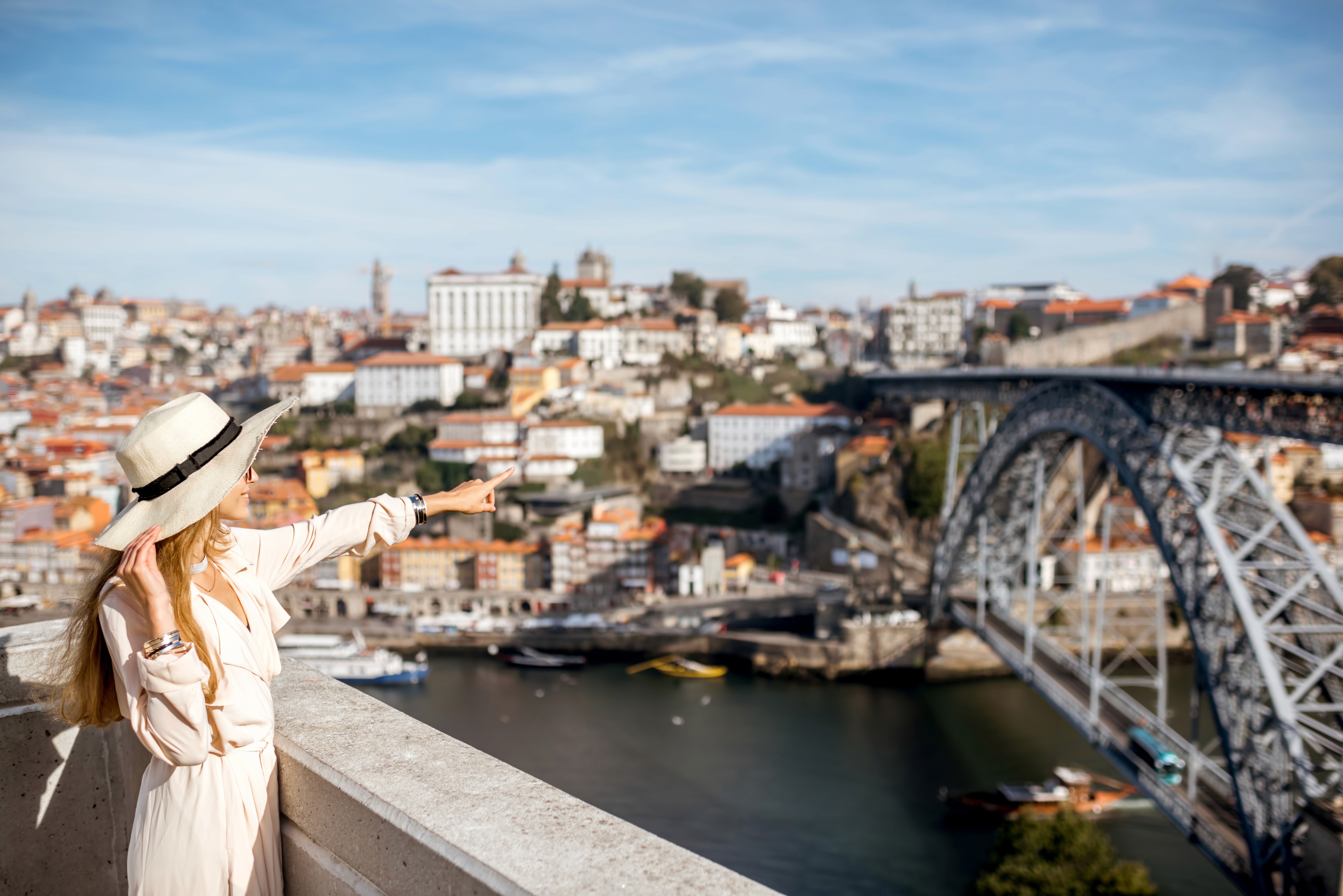 Imagem mostra uma mulher de conjunto e chapéu brancos, cabelos loiros, apontando em direção à ponte D. Pedro I, com a cidade do Porto ao fundo.