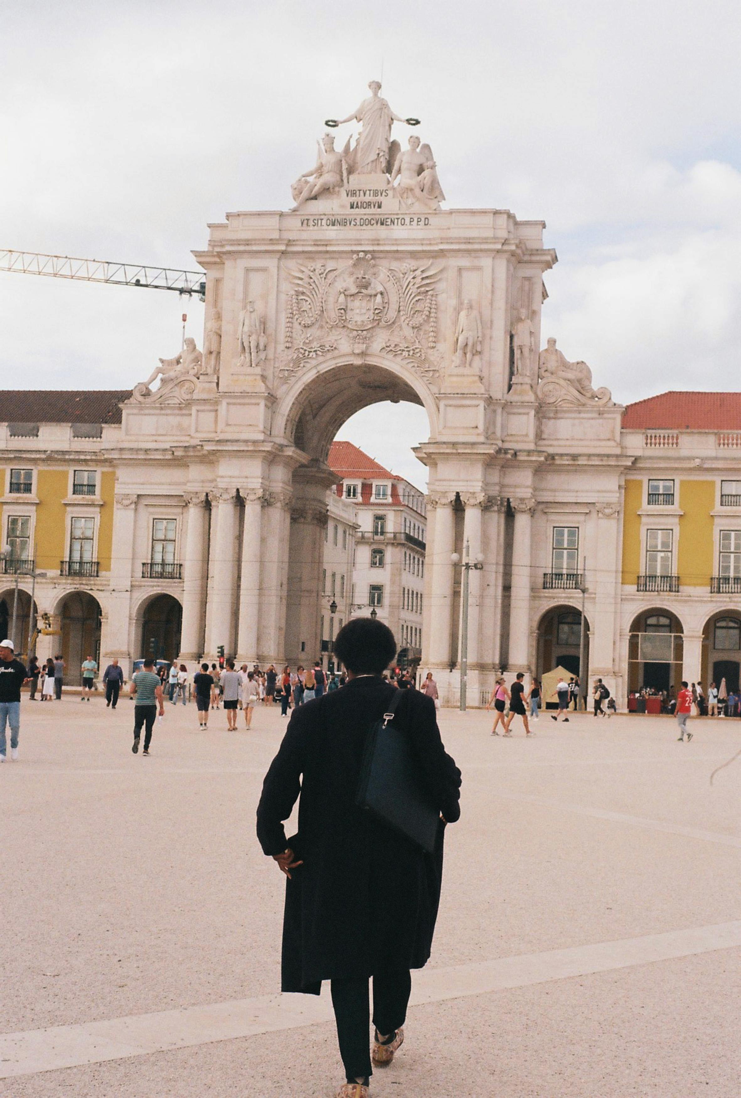 Homem, trajando sobretudo e bolsa preta, andando em direção à Praça do Comércio em Lisboa, Portugal.