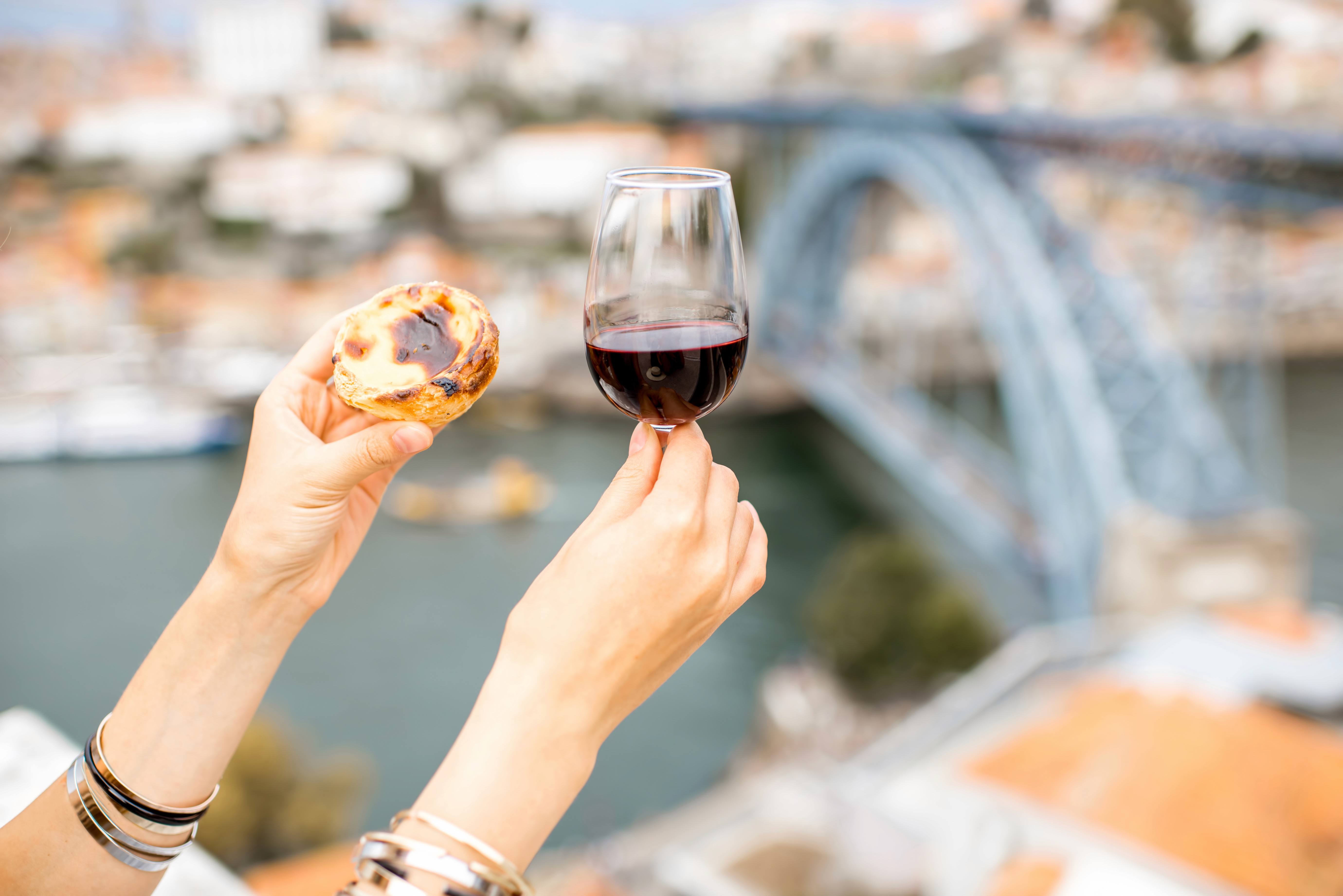Na imagem, uma mão segura uma taça de vinho tinto, enquanto a outra segura um pastel de nata,com o Rio Douro, que passa na cidade do Porto, ao fundo desfocado.