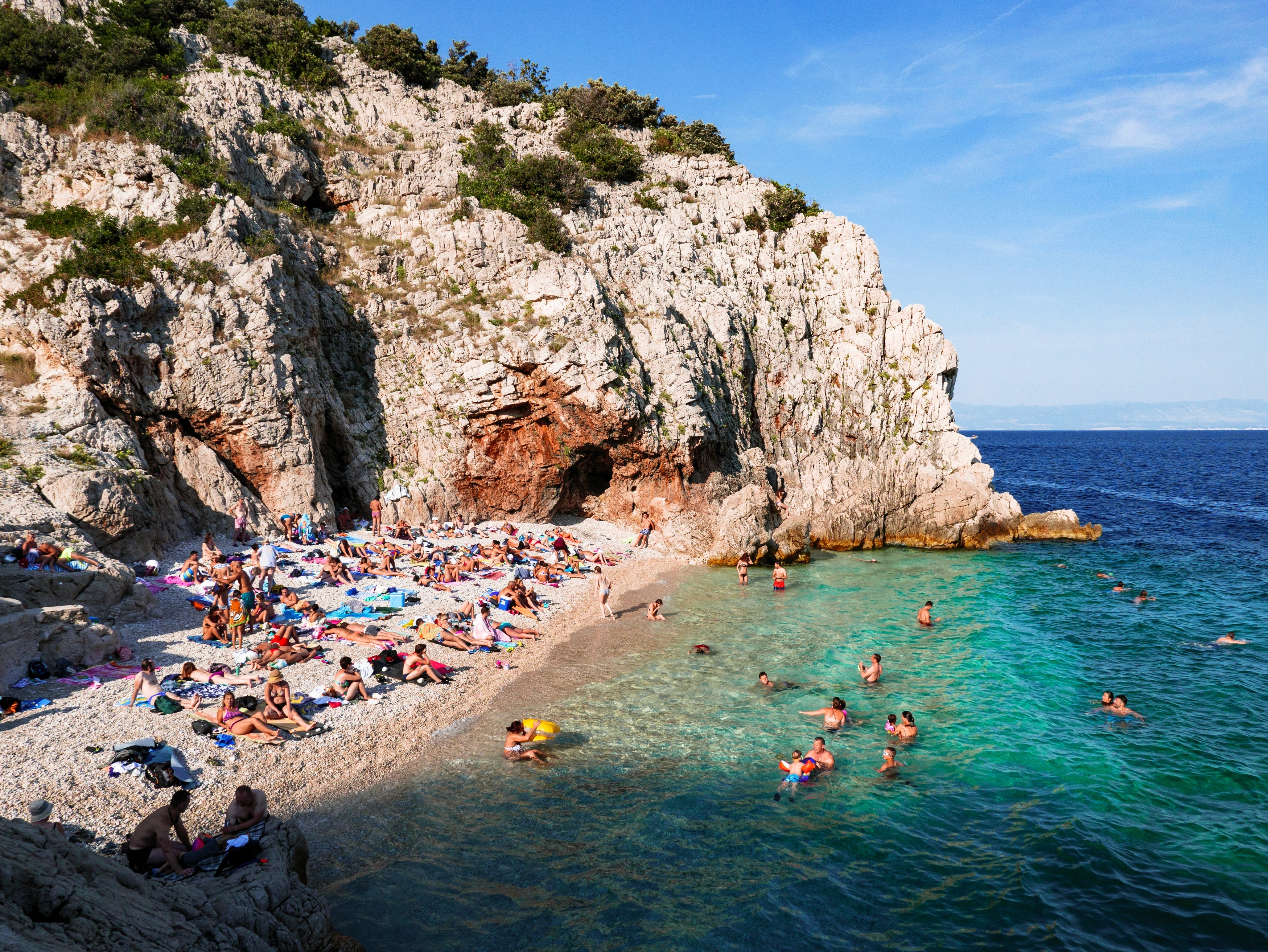 Diversas pessoas se divertindo numa praia, com uma grande falésia atrás. Algumas pessoas tomam banho na água azul e límpida.