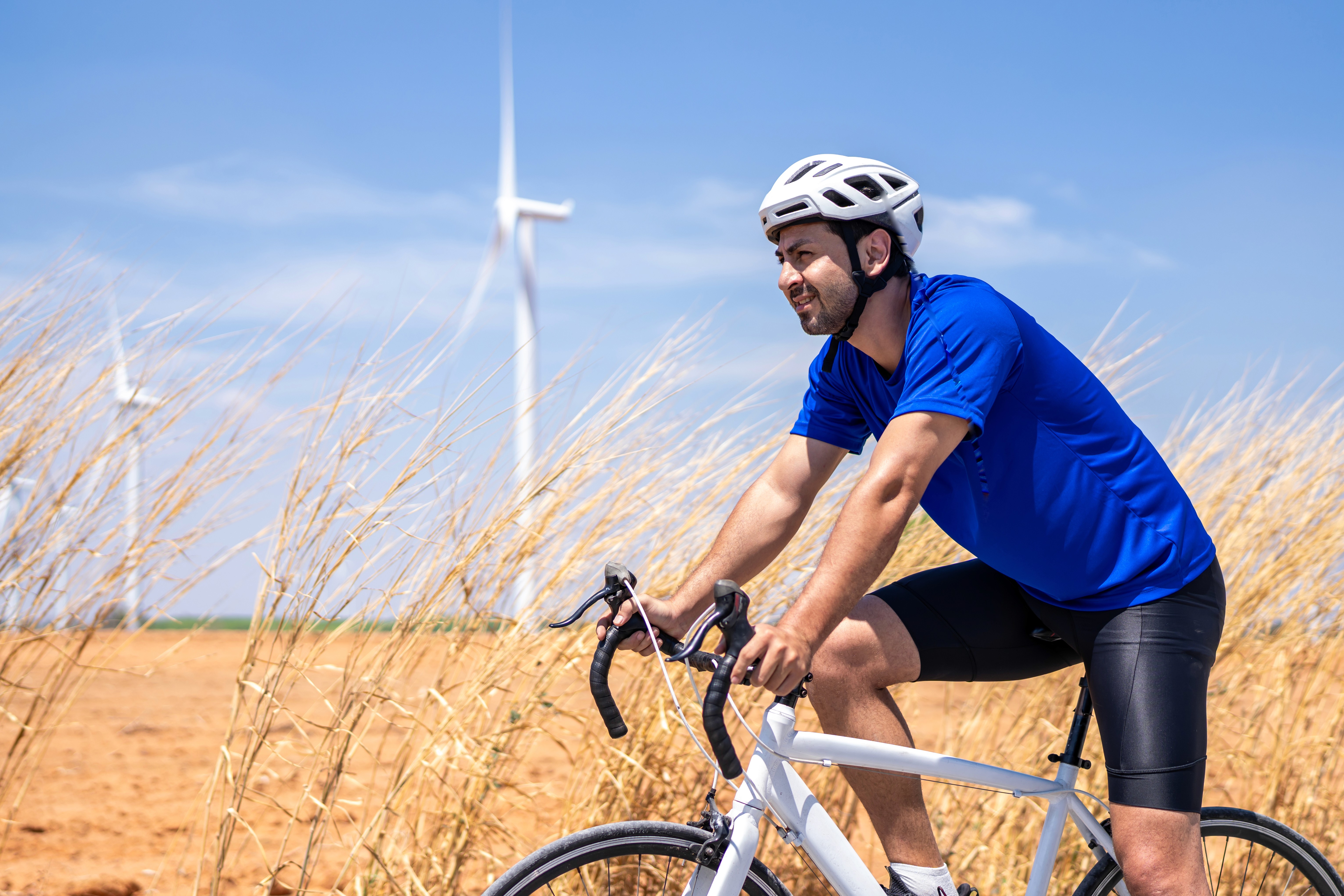 Um ciclista homem, branco, de camiseta azul e bermuda de lycra escura, pedala com um campo de centeio ao fundo. 