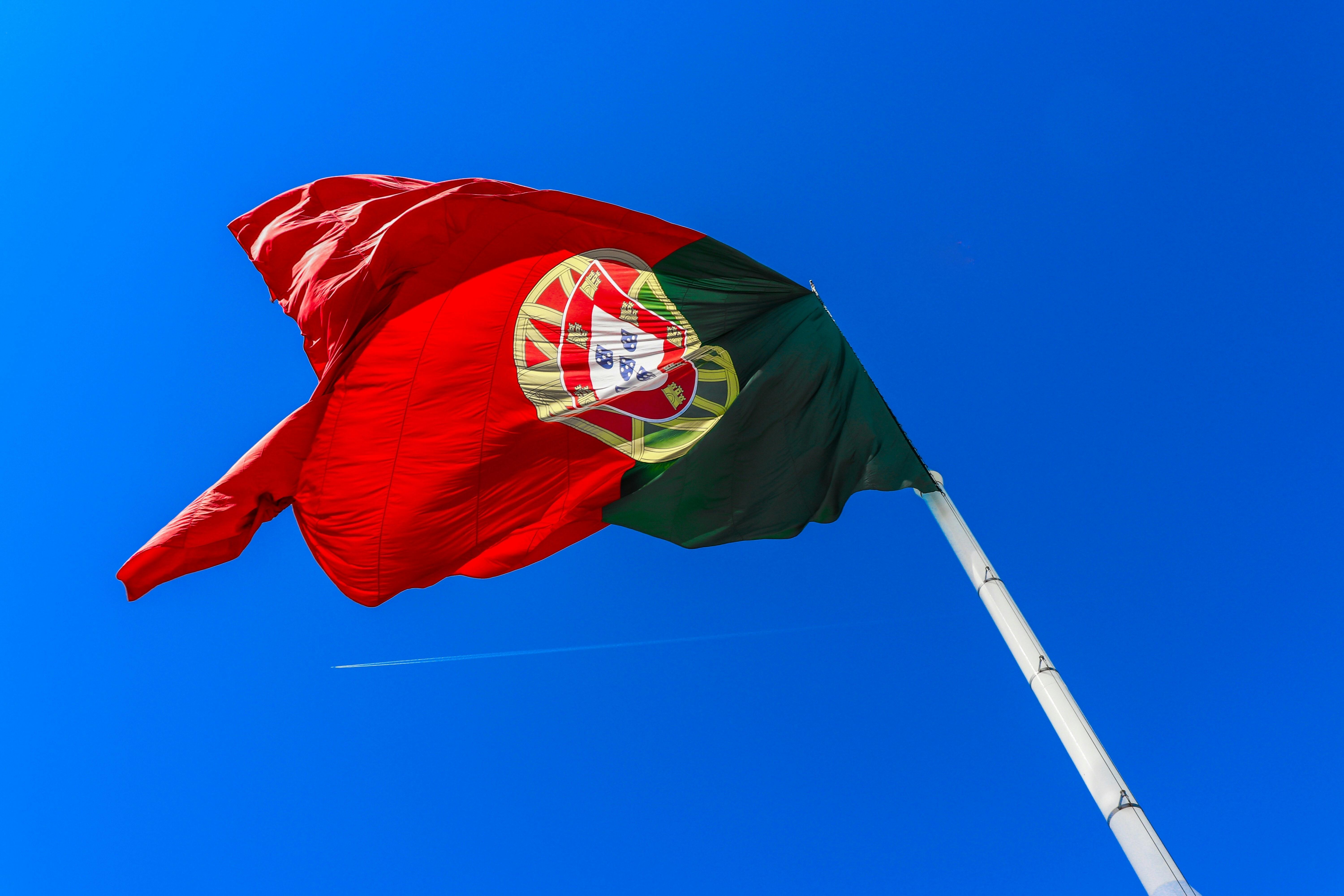 Imagem mostra a bandeira portuguesa estendida em um mastro, em ângulo contra-plongée, com um vasto céu azul ao fundo. 
