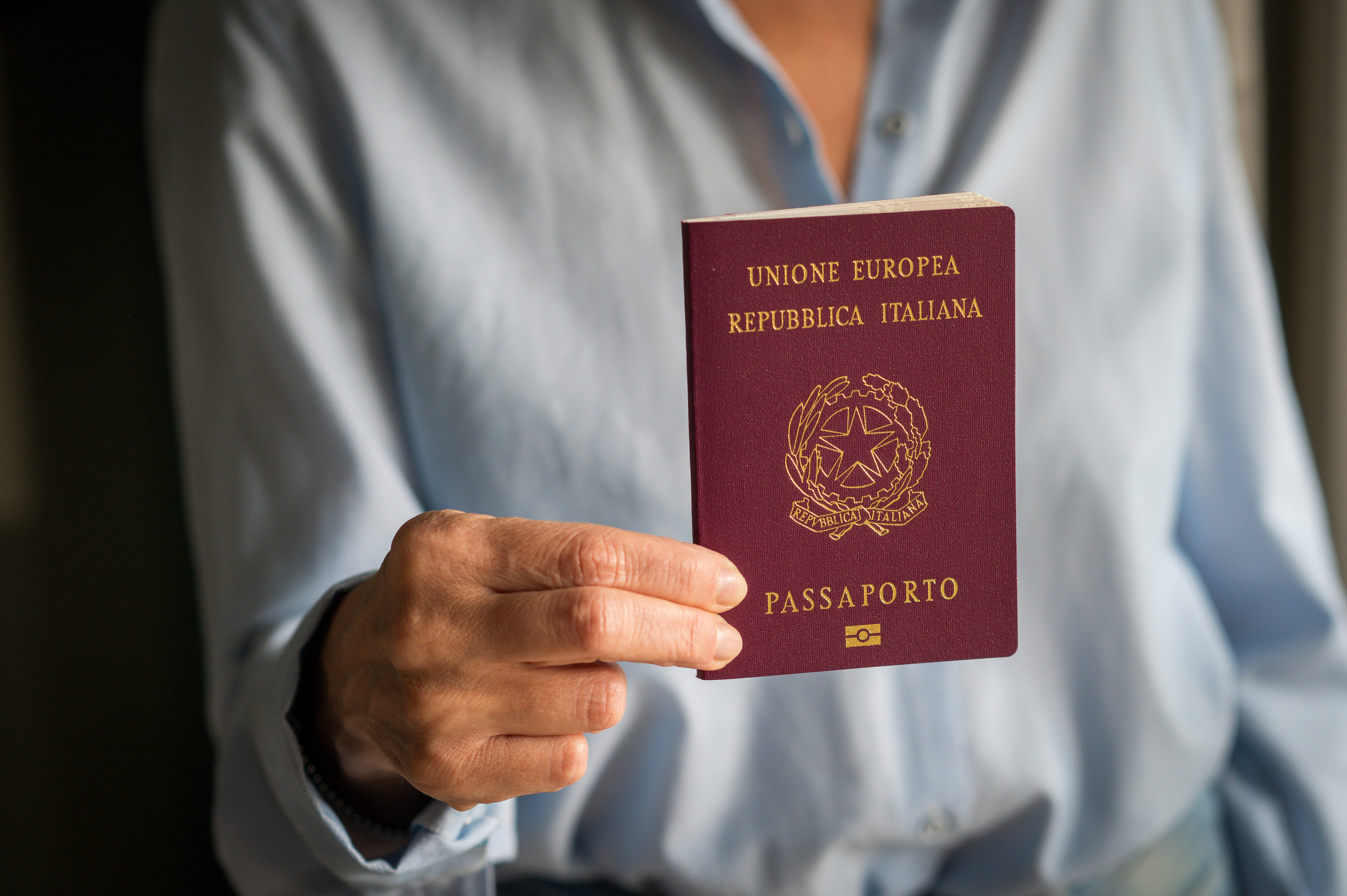 Mulher de camisa casual azulada segurando um passaporte italiano, com o fundo levemente desfocado