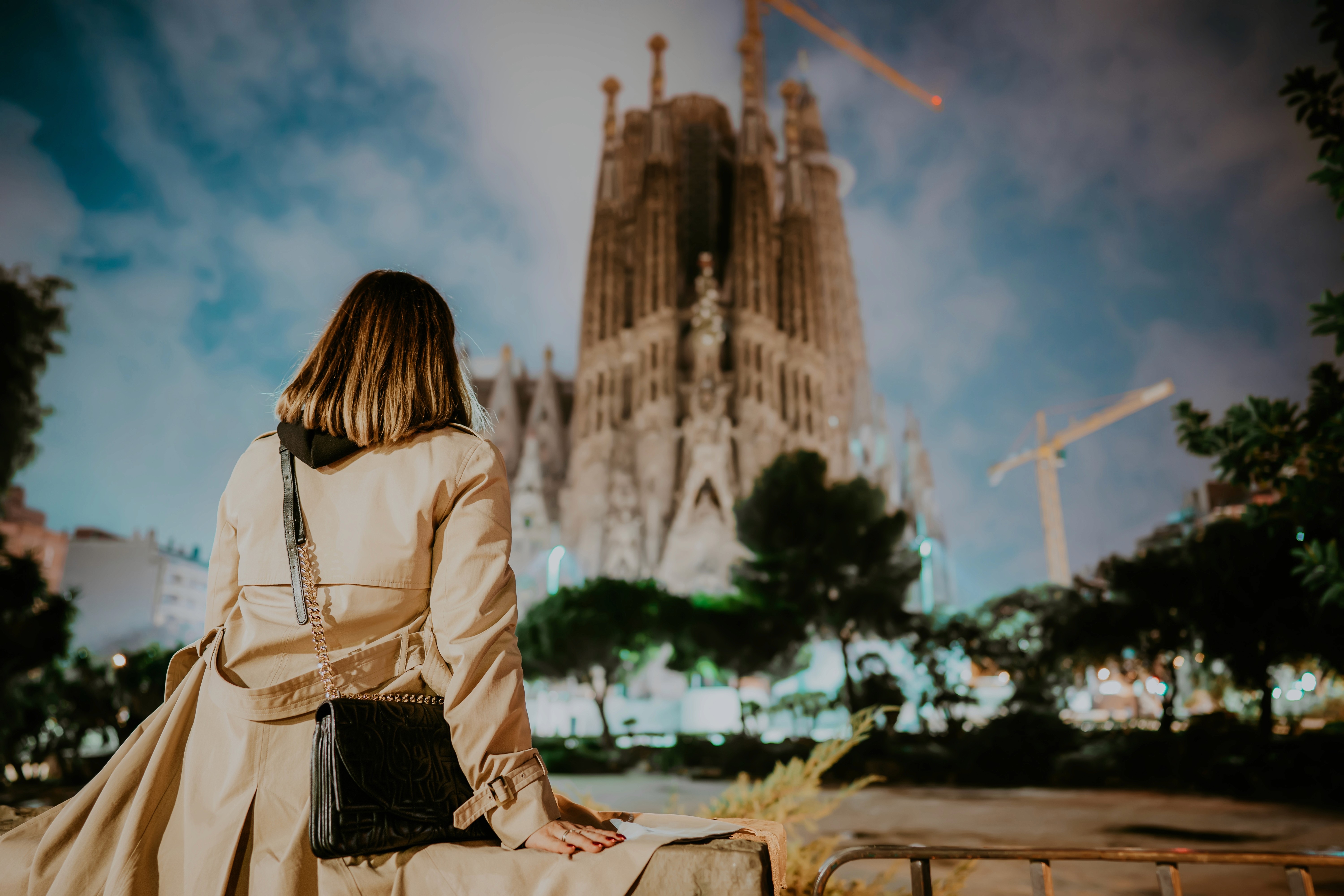 Mulher sentada, de cabelos curtos, sobretudo e bolsa lateral, sentada enquanto observa a Sagrada Família, símbolo de Barcelona e obra-prima de Antoni Gauldí, à noite.