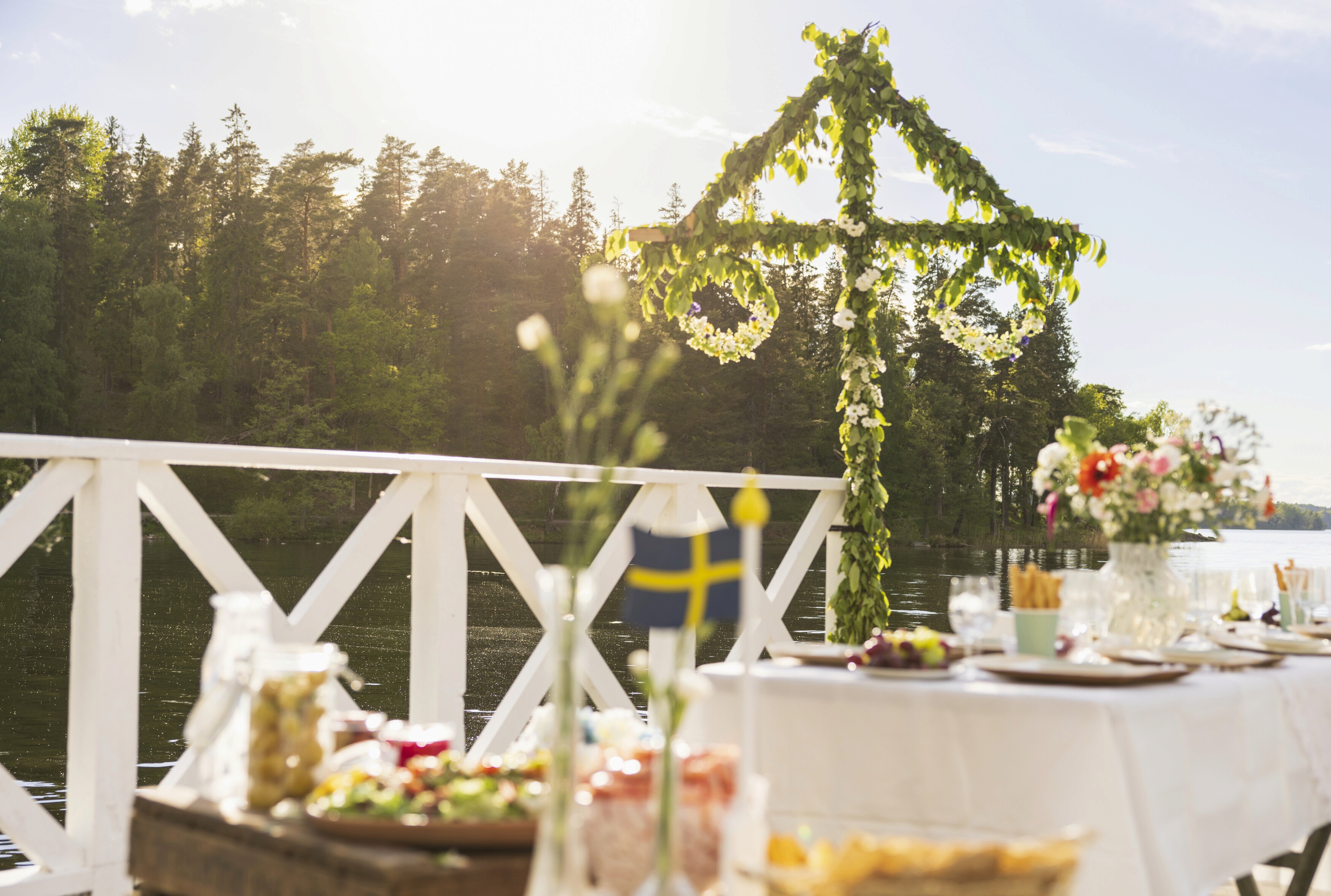 Na foto, uma mesa posta com diversas comidas em um campo verde com diversas árvores e o sol no topo da imagem, iluminando tudo com seus raios. Ao fundo, um majstången, um mastro do verão que é símbolo das festividades.