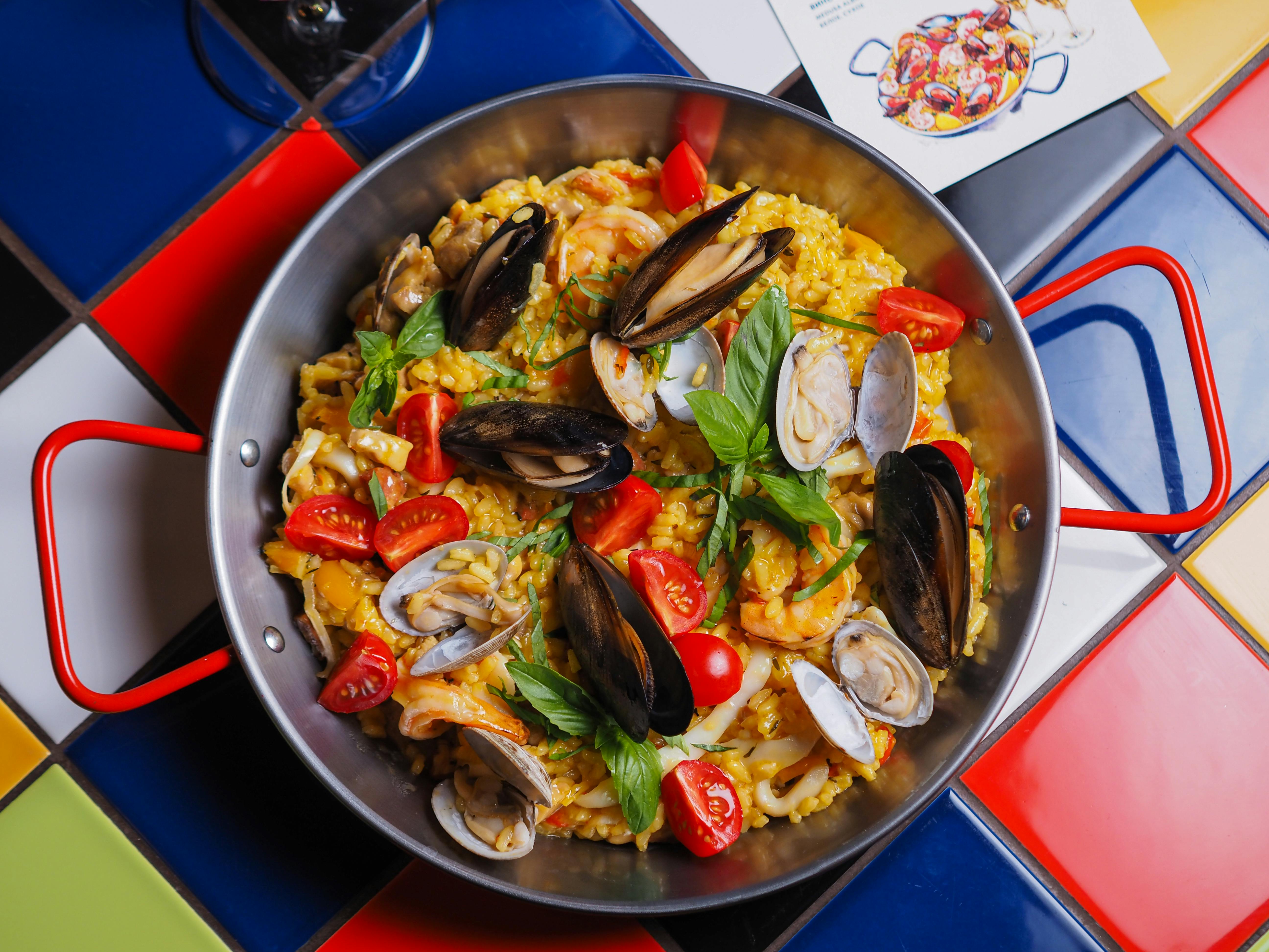 Visão de cima de uma panela de Paella, tradicional prato espanhol feito com arroz temperado com molho de açafrão e frutos do mar, sob uma mesa colorida.