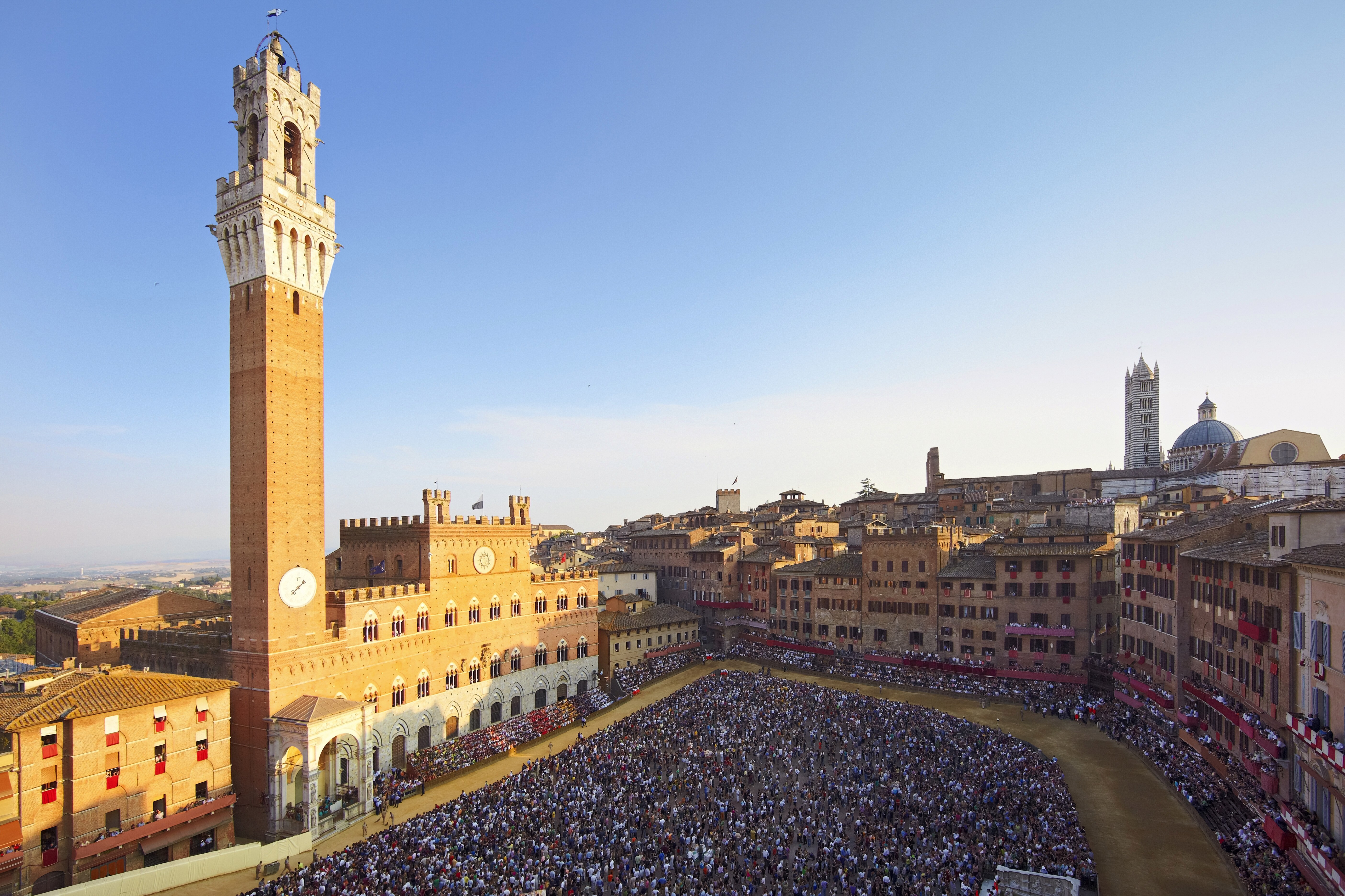 Vista ampla da Piazza del Campo, em Siena. Principal ponto da cidade, é onde ocorre o Palio di Siena todos os anos. Várias pessoas, uma multidão se reúne, com uma parte do público disperso em arquibancadas e o restante, ao centro da pista