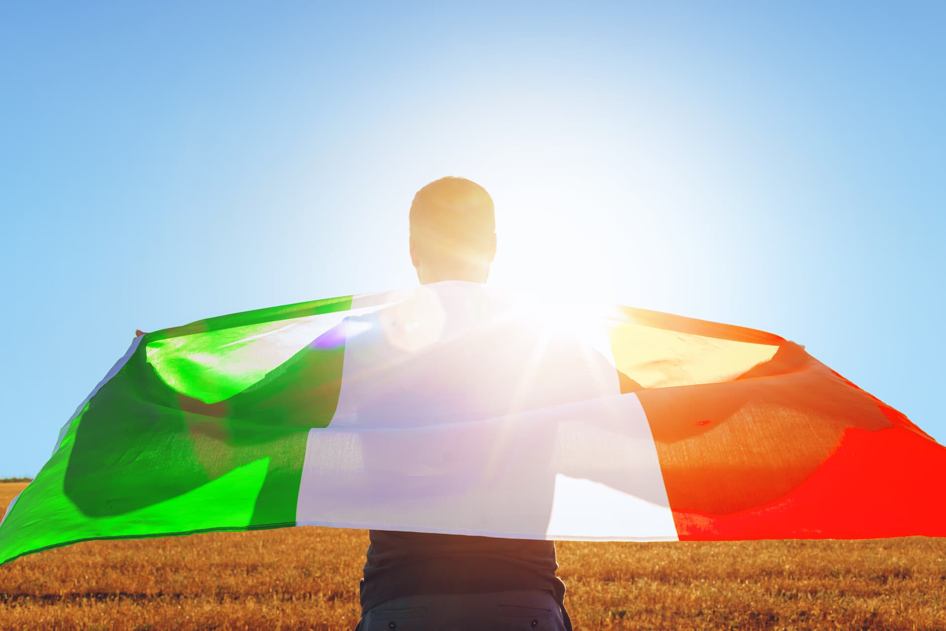 Homem carrega bandeira da Itália estendida em suas costas, com o pôr do sol ao fundo brilhando fortemente. O que representa o Dia da Unidade Nacional, da Constituição, do Hino e da Bandeira na Itália
