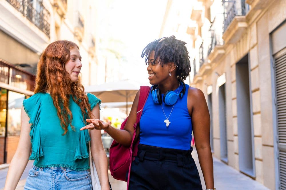 Duas mulheres, uma negra com curtas tranças soltas, e outra branca, de cabelo ondulado ruivo, conversam enquanto andam por uma rua.