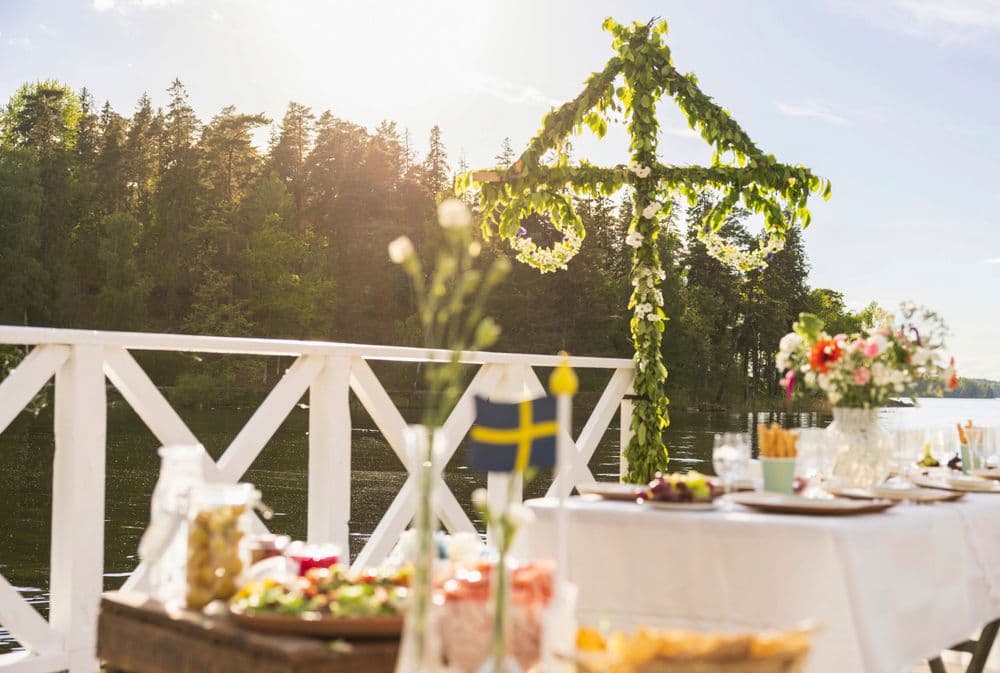 Na foto, uma mesa posta com diversas comidas em um campo verde com diversas árvores e o sol no topo da imagem, iluminando tudo com seus raios. Ao fund