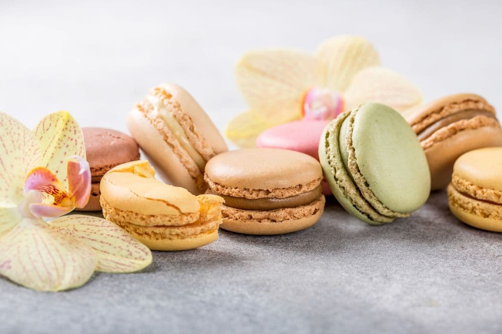 Diversos macarones sob uma toalha de tom acinzentando, com algumas fores a decorando