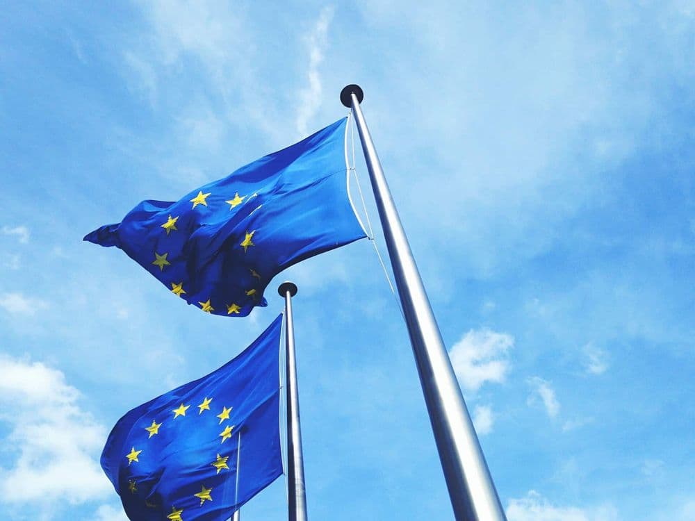 Duas bandeiras da União Europeia hasteadas e balançando ao vento, com o céu azul com poucas nuvens ao fundo.